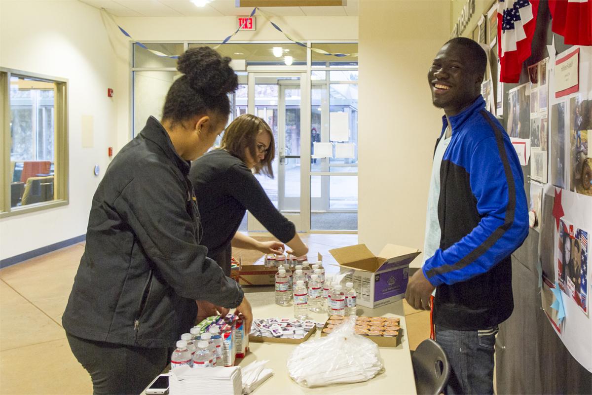 A smiling male student and two female students distribute snacks in the Student Center